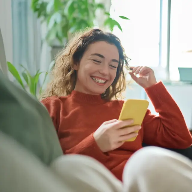 Happy relaxed young woman sitting on couch using mobile phone